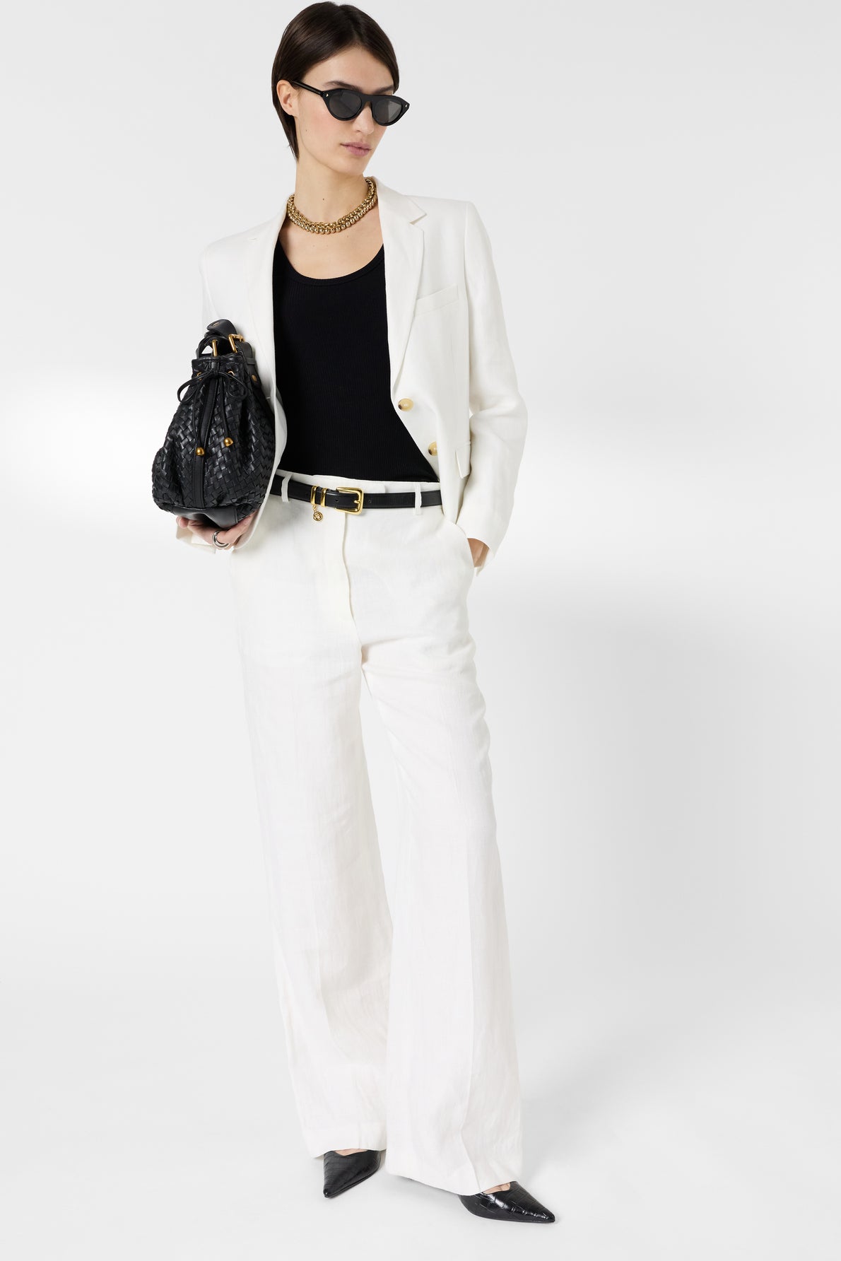 Woman in white suit with black top, black pointed shoes, holding a black handbag, wearing sunglasses and a chunky gold necklace, standing against a plain white background.