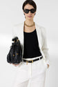 Woman in a white suit with a black top, wearing sunglasses and a gold necklace, holds a black woven handbag and stands confidently against a plain white background.