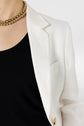 A person wearing a white blazer over a black top, accessorized with a chunky gold necklace. Only the upper torso is visible against a plain background.