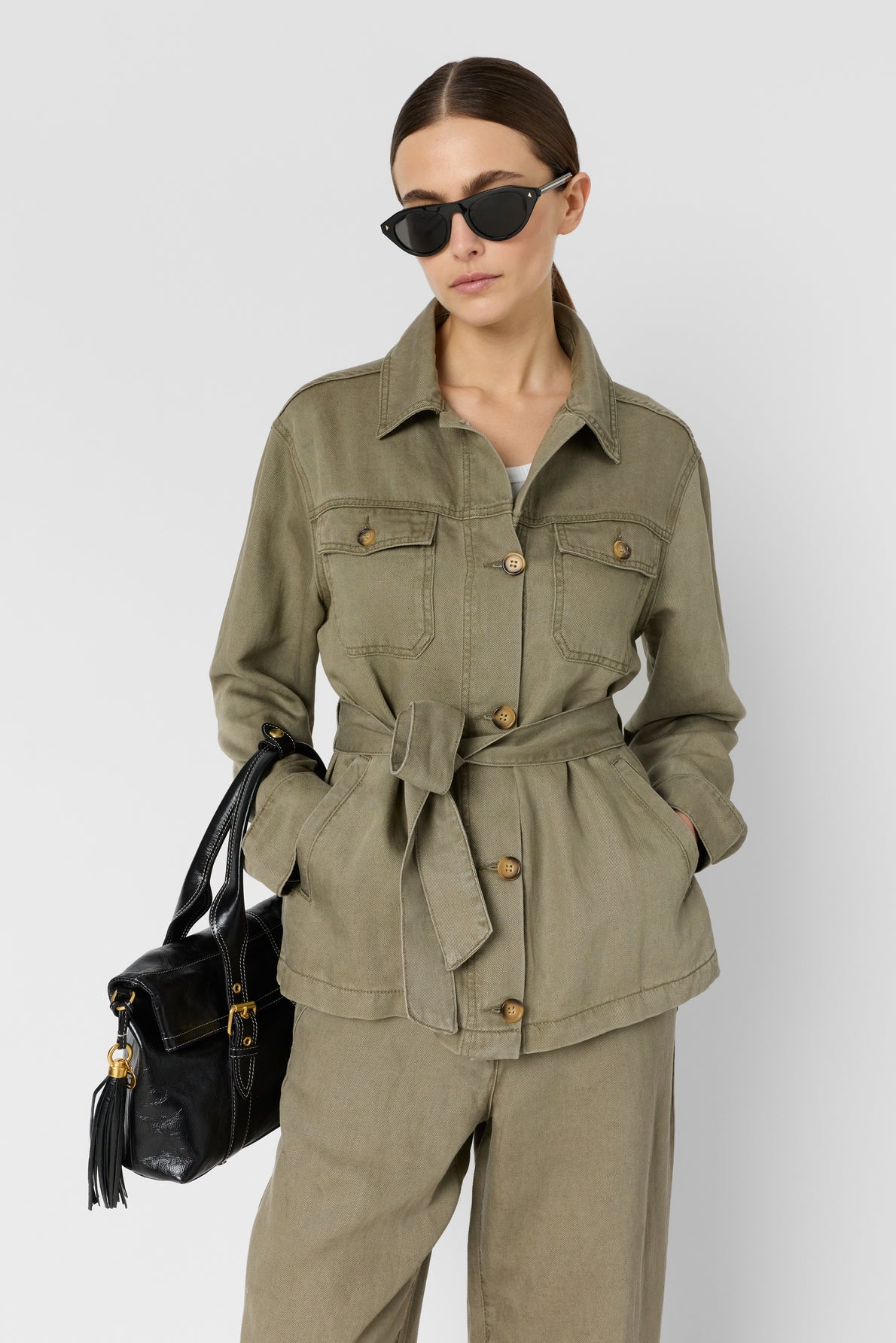 Woman wearing an olive green lyocell lin veste saharienne, paired with matching pants, holding a black handbag and wearing black sunglasses. She stands against a plain light background.