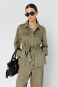 Woman wearing an olive green lyocell lin veste saharienne, paired with matching pants, holding a black handbag and wearing black sunglasses. She stands against a plain light background.