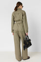 A woman stands facing away, wearing a veste saharienne in olive green with a belted waist and matching wide-leg pants crafted from lyocell lin. She holds a black handbag and wears black sandals and sunglasses, her hair tied back in a low ponytail.