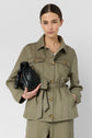 A woman with brown hair in a low bun wears a veste saharienne olive green belted jacket crafted from lyocell lin and holds a black handbag. She stands against a plain light background, looking directly at the camera with a neutral expression.
