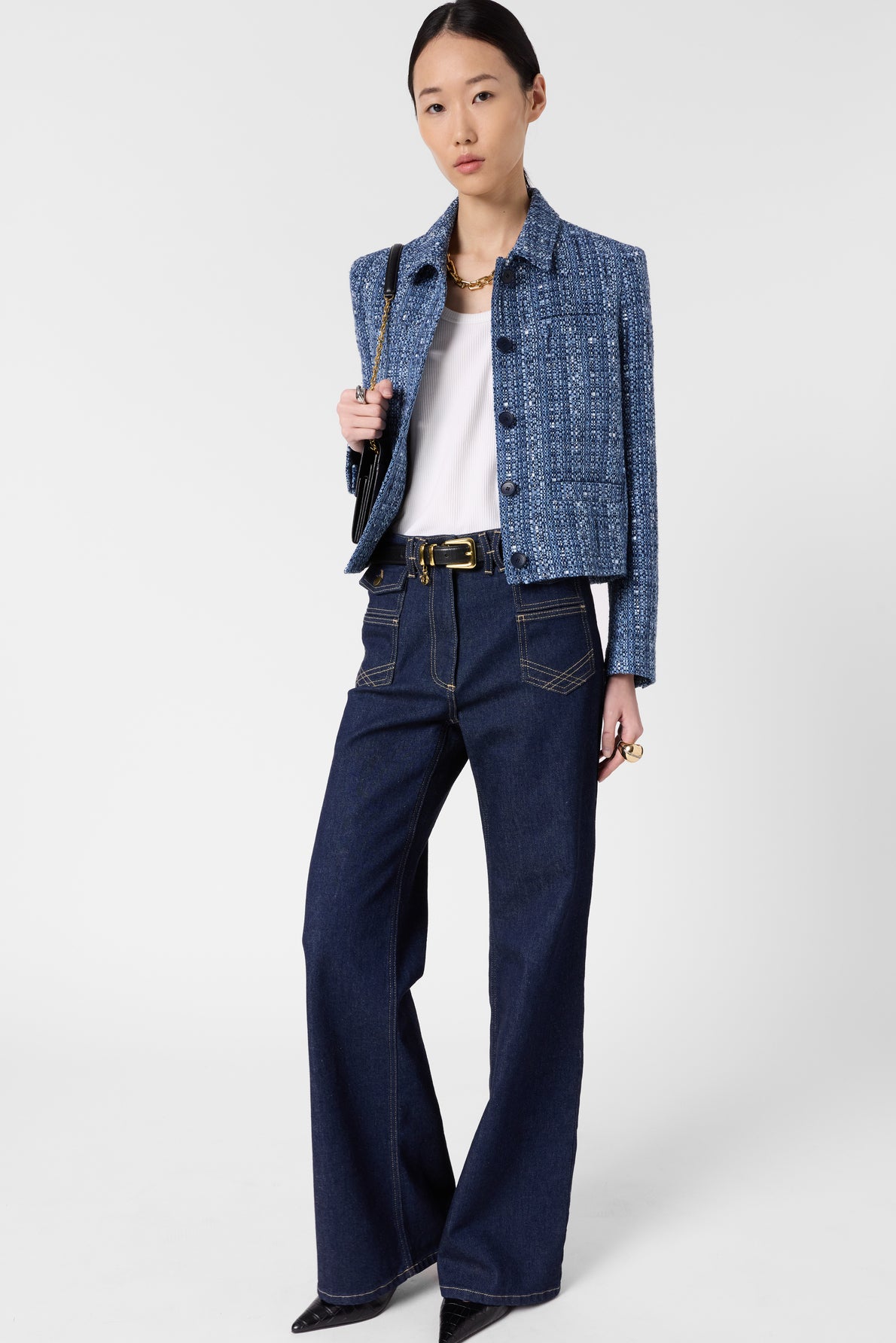 Woman standing against white background, wearing a blue textured jacket, white top, dark high-waisted wide-leg jeans with large pockets, black pointed shoes, and holding a black bag on her shoulder.