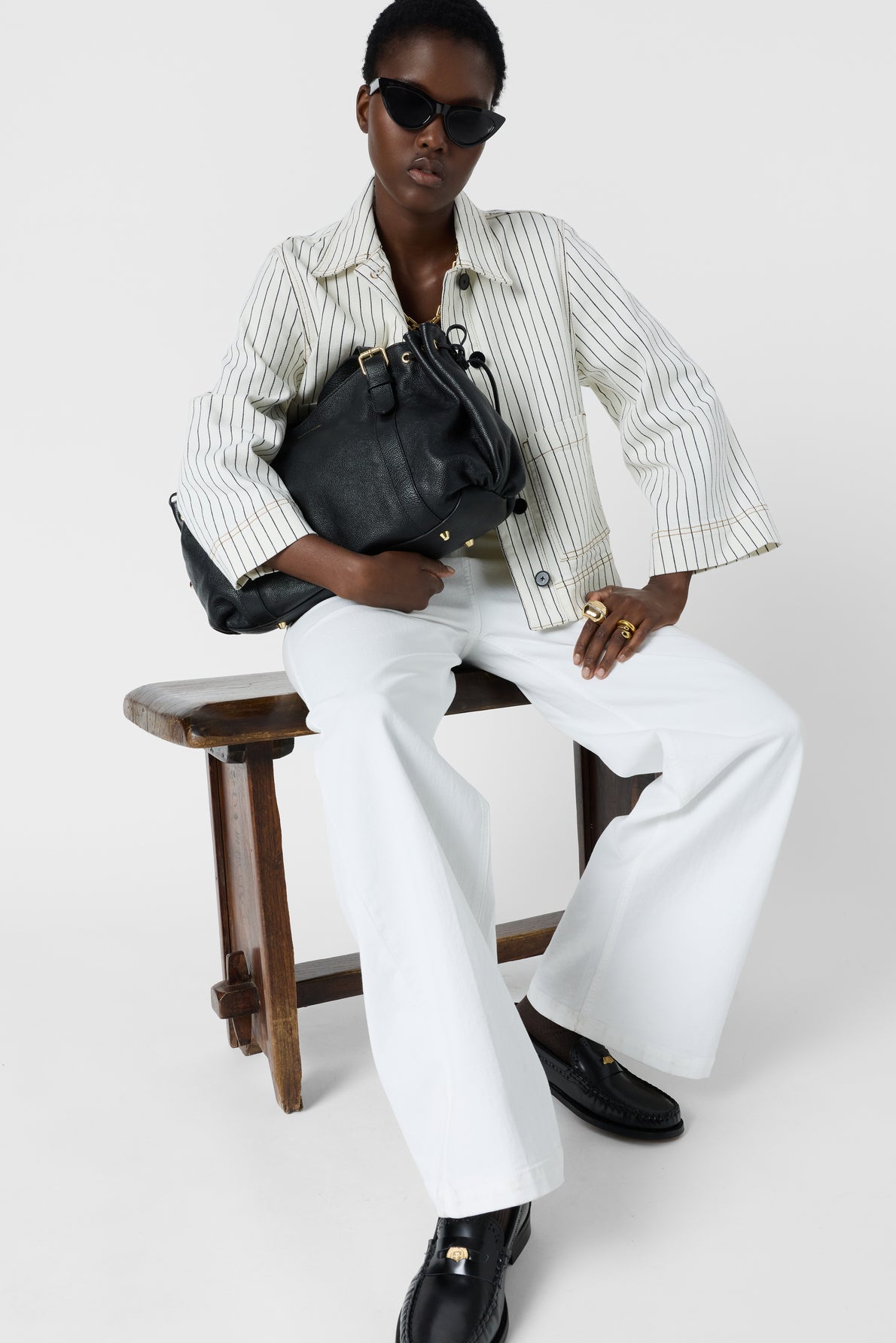 A person wearing sunglasses, a veste rayée femme—a stylish striped white jacket—white pants, and black shoes sits on a wooden bench, holding a large black bag across their lap.