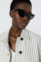 A person wearing black cat-eye sunglasses, a gold chain necklace, a white shirt, and a veste rayée femme—specifically a white jacket with black vertical stripes—stands against a plain background.