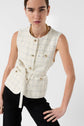 Woman in a sleeveless cream plaid vest with gold buttons and a belt, paired with black pants, leans slightly forward against a plain white background.