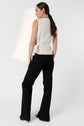 Woman with long brown hair in ponytail, wearing a sleeveless, white plaid top and black pants, stands facing away with one hand in her pocket against a plain light background.