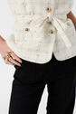 A person wearing a cream plaid buttoned jacket tied at the waist, black pants, and gold rings. Only the torso and one hand are visible.