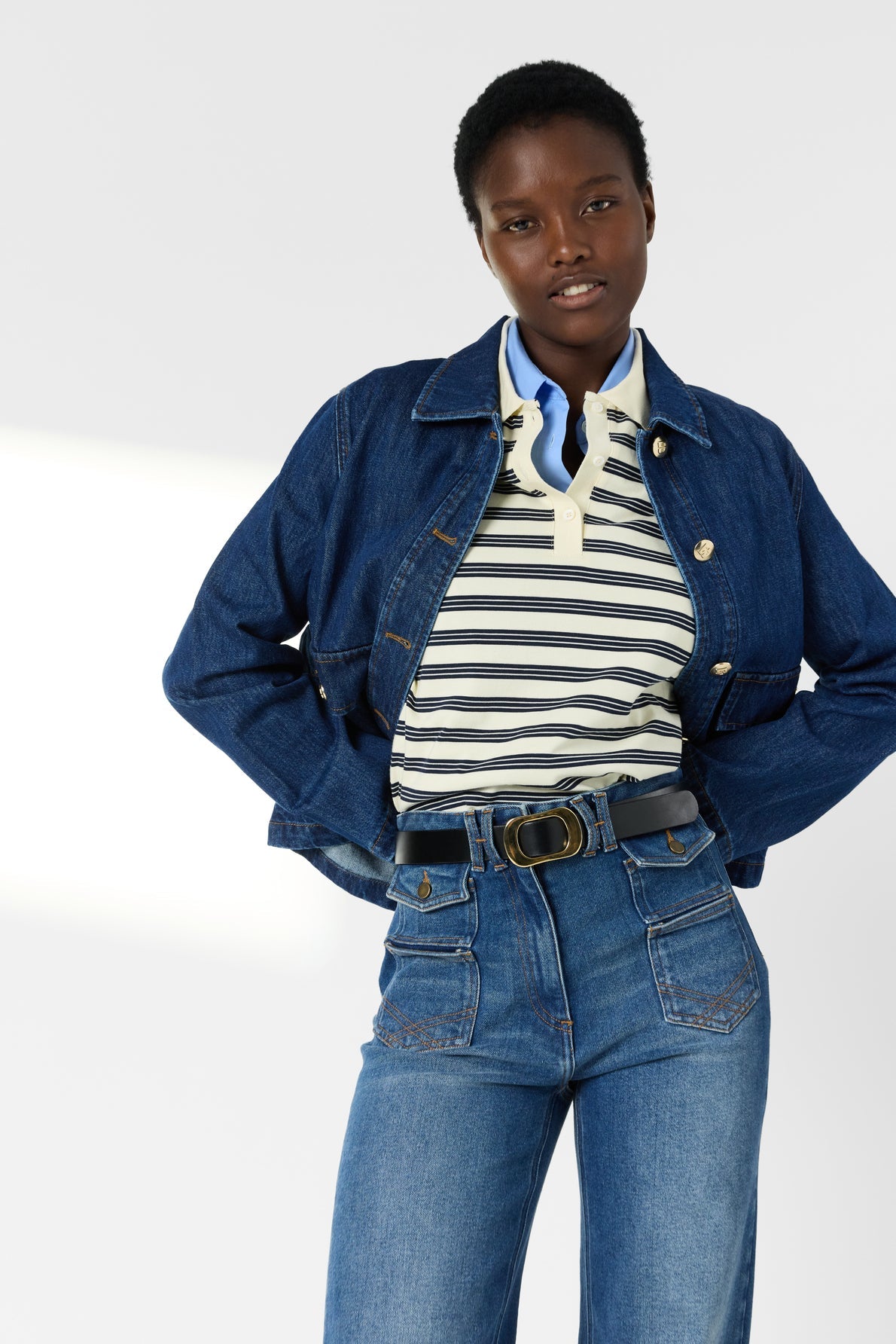 A person wearing a veste courte en denim, striped polo shirt, and high-waisted jeans with front pockets stands with hands on hips, looking at the camera.