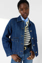 A person wearing a veste en denim, styled as a dark denim jacket over a striped shirt and blue collared shirt, with blue jeans and gold rings, poses against a light background.
