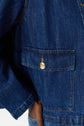 Close-up of a denim jacket pocket with a gold button. The stitching and texture of the blue denim fabric are clearly visible, highlighting the classic style of this veste droite femme. A person’s arm is partially seen resting on their waist, emphasizing the structured fit of this elegant veste en denim.