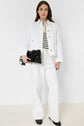 Woman wearing a veste en jean blanc and wide-leg white pants in coton stretch, paired with a striped shirt and black shoes. She holds a black clutch bag and stands against a plain light background.
