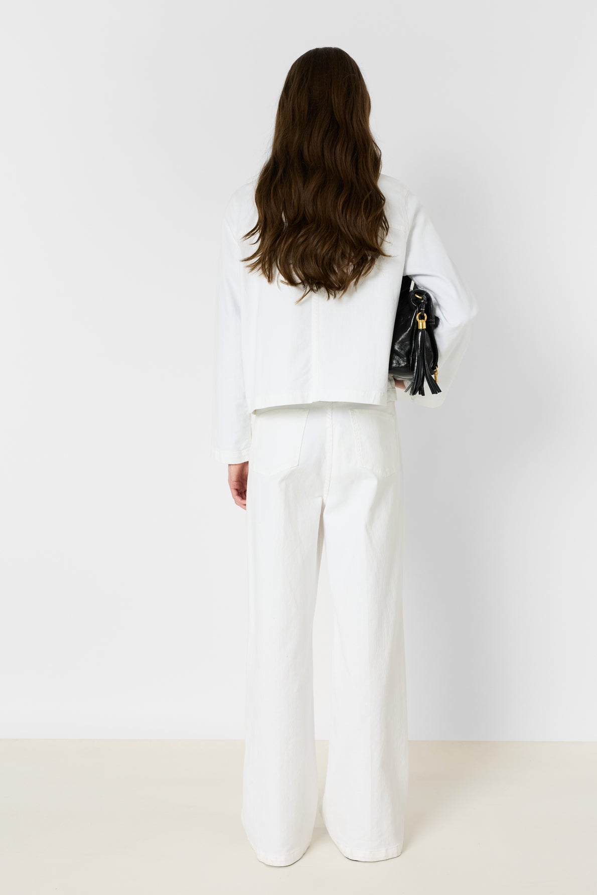 Woman with long wavy brown hair stands facing away, wearing a white coton stretch long-sleeve top and wide-leg pants, holding a black handbag under her left arm against a plain white background.