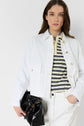 A woman in a veste courte blanche and pants with a striped shirt holds a black handbag. She has long brown hair and stands against a plain white background, one hand casually tucked in her pocket. The outfit features coton stretch for effortless comfort and style.