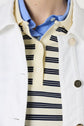 A close-up of a person wearing a blue collared shirt under a cream and navy striped polo, topped with a veste en jean blanc. Only the neck and upper chest are visible, highlighting the crisp layers under the short white jacket crafted in coton stretch.