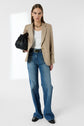 A woman in a beige blazer, white top, and blue jeans stands holding a black textured handbag. She wears black shoes and a gold necklace, with a neutral expression against a plain white background.