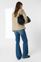 A person stands facing away, wearing a beige blazer, blue jeans, and black shoes, holding a large black textured handbag under one arm against a plain white background.