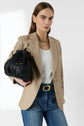 A woman wearing a beige blazer, white ribbed top, blue jeans, and gold necklace holds a black woven handbag. She stands against a plain white background, looking confidently at the camera.