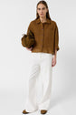 A person stands against a white background wearing a brown button-up shirt, white wide-leg pants, brown sandals, and holding a brown bag.