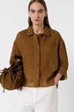 A woman with straight brown hair wears a brown button-up shirt with a chest pocket, white pants, and holds a brown drawstring bag against a white background.