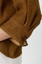 A close-up of a persons arm wearing a brown textured, mesh-like long sleeve with a buttoned cuff, paired with white pants.