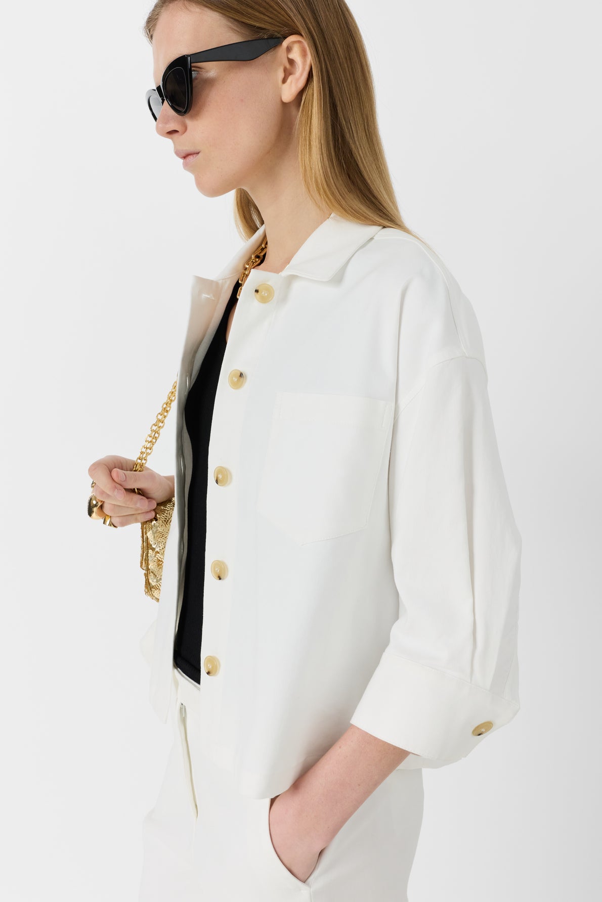 Woman with long blonde hair wearing black sunglasses, a white button-up jacket, and white trousers, holding a gold chain purse and dressed in a black top, poses against a plain white background.