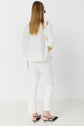 A person with long light brown hair, wearing a loose white top, white pants, and black slingback shoes, stands facing away from the camera against a plain white background.