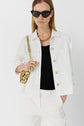 A woman in white pants and a white buttoned shirt over a black top, wearing sunglasses and a gold necklace, holds a gold bag on her shoulder.