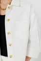 A person wearing a white button-up shirt with beige buttons, a black top underneath, a gold chain necklace, and white pants. The image is cropped to show only the upper torso.