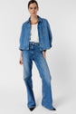 A person wearing a loose denim jacket, white top, and wide-leg blue jeans with black pointed shoes stands against a plain white background.