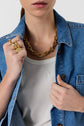 A close-up shot of a person wearing a white tank top under a denim jacket, adorned with a chunky gold chain necklace and gold rings on their fingers.