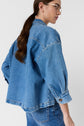 A person with long brown hair, wearing glasses, a blue denim jacket, and matching blue jeans stands against a plain light background, facing slightly away from the camera.