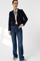 A person stands against a white background wearing a navy blazer, white blouse with a patterned bow, blue jeans, and white shoes.