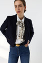 A woman wearing a dark blazer, white blouse with a patterned bow, blue jeans, and a gold-buckled belt stands with one hand on her hip against a plain light background.