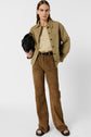 Woman standing against a white background, wearing a veste utilitaire kaki, cream top, brown suede pants, and black sandals, holding a black bag under her arm. She has straight brown hair and a neutral expression.