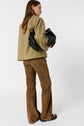 A woman with long brown hair, wearing a veste utilitaire kaki and brown suede pants, stands facing away while holding a black handbag under her arm. She is also wearing black sandals.