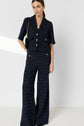 A person stands against a plain background wearing a navy blue plaid short-sleeve jacket with gold buttons and matching high-waisted wide-leg pants, accessorized with a black shoulder bag and gold jewelry.