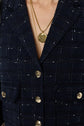 A close-up of a person wearing a navy plaid textured blazer with gold buttons and two gold necklaces featuring coin pendants.