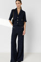 Woman wearing a navy blue plaid pantsuit with gold buttons, short sleeves, and wide-leg pants. She has a black shoulder bag and gold jewelry, standing against a plain light background.