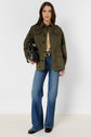 Woman standing against a white background wearing a veste militaire kaki, tan shirt, blue flared jeans, black shoes, and holding a black handbag.
