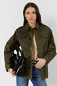 A woman wearing a veste utilitaire femme—a khaki green button-up jacket—over a tan shirt, blue jeans, and a black belt, holds a black handbag and looks at the camera against a plain white background.