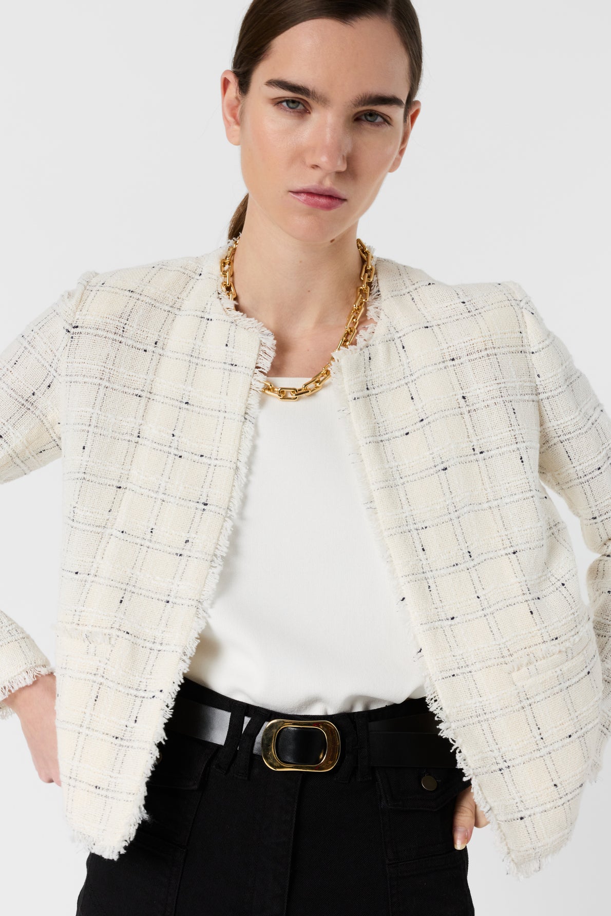 Woman wearing a textured white plaid veste écru over a white top, black pants with a belt, and a chunky gold chain necklace, standing with hands on hips against a plain white background.