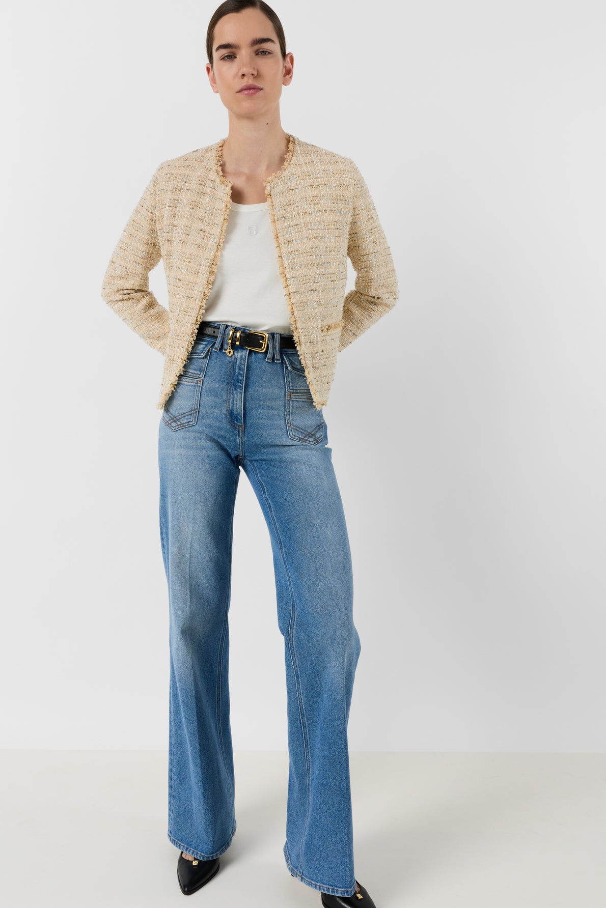 A person stands against a white background wearing a cream veste tweed femme, white top, blue high-waisted jeans with a black belt, and black pointed shoes, looking confidently at the camera.