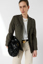 A person in a dark olive veste saharienne blazer, white shirt, and light pants holds a large black leather bag. They wear a gold necklace and a neutral expression, standing against a plain light background.