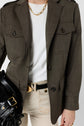 A person wearing a dark olive veste saharienne blazer, white shirt, beige pants, black belt, and a chunky gold necklace, holding a black leather bag with gold hardware.