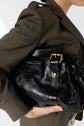 A person in a veste chic brown blazer holds a shiny black leather handbag under their arm, showing a close-up of the bag’s buckle and textured surface.