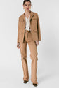 A woman stands wearing a tan utility jacket reminiscent of a veste saharienne, matching flare pants with frayed seams, a white blouse, brown shoes, gold hoop earrings, and a belt. She poses against a plain white background.