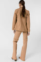 Woman with long brown hair in a ponytail wearing a veste coton beige and matching flared pants, standing and facing away on a plain light gray background.