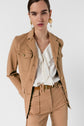 A woman wearing a tan veste saharienne and matching pants with a white ruffled blouse stands against a plain white background, looking confidently at the camera. She has gold earrings and her hands are behind her back.
